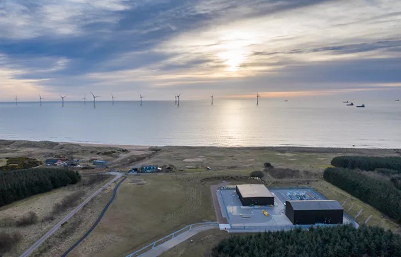The ocean with boats and wind turbines next to land with fields, trees, and industrial buildings.