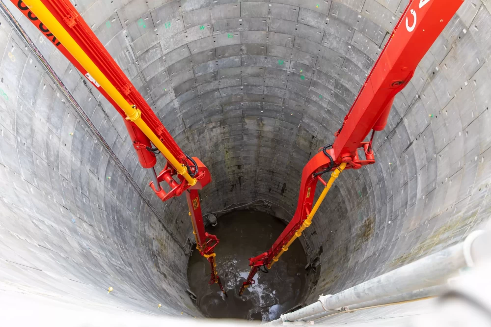 Red and yellow construction vehicles pouring concrete into a large grey cylinder.