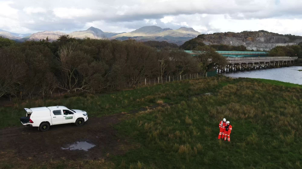 Three people, one in a white Murphy vehicle and two wearing safety equipment in a field with a bridge over water in the background.