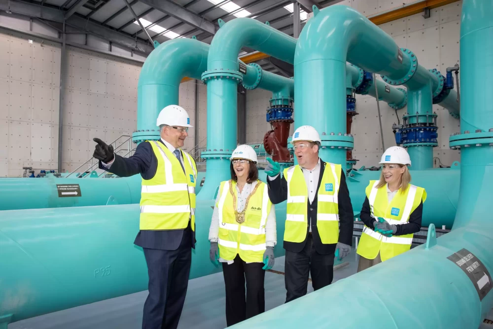 Four people in safety uniform stood in a room smiling surrounded by large blue pipes.