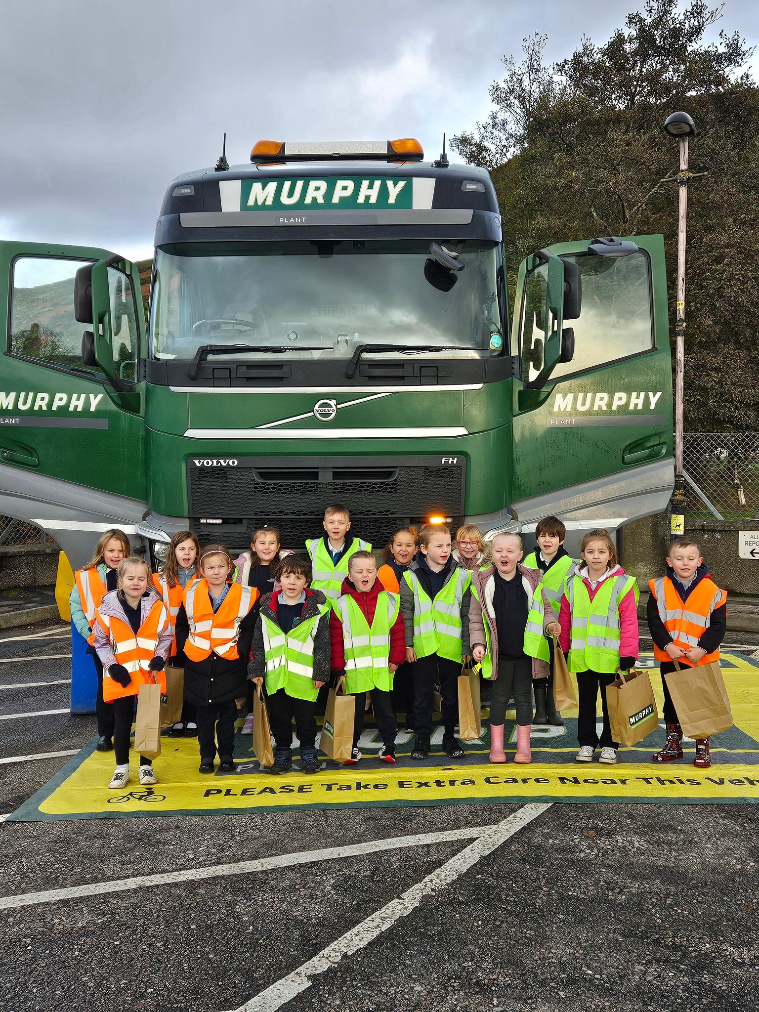 A group of children wearing high visibility jackets carrying Murphy branded brown paper bags stood in front of a green Murphy lorry with the doors open.
