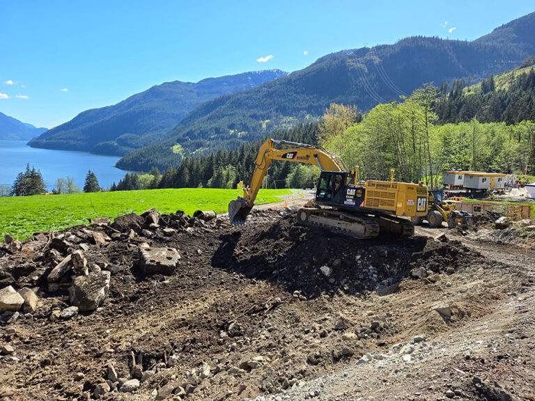 A gas pipeline construction site with a yellow construction vehicle and mountains, trees and water in the background.