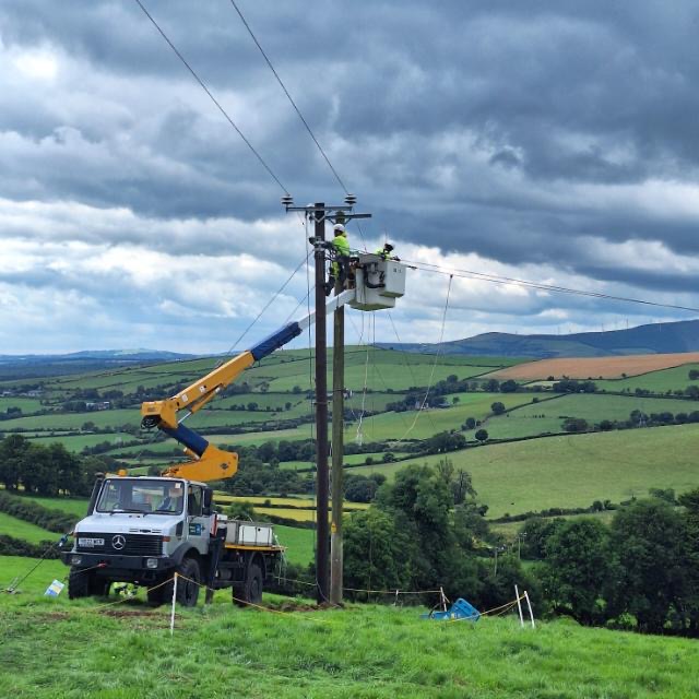 A person wearing safety equipment working on an overhead line in a field.