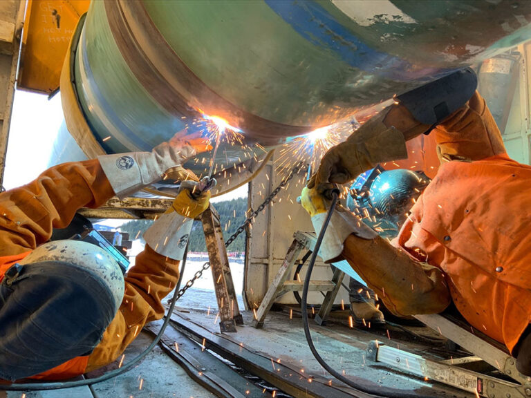Two workers wearing safety equipment conducting work on a large metal pipe.