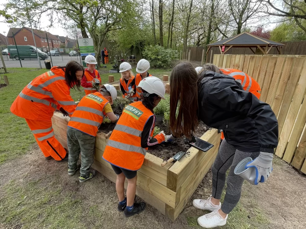 Two adults and a group of children wearing Murphy branded high-visibility clothing and white helmets working on a garden.