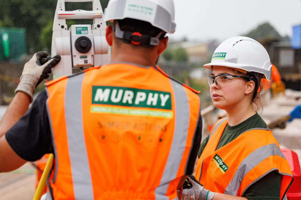 Two people in Murphy high-visibility jackets and helmets on a construction site operating a piece of equipment.