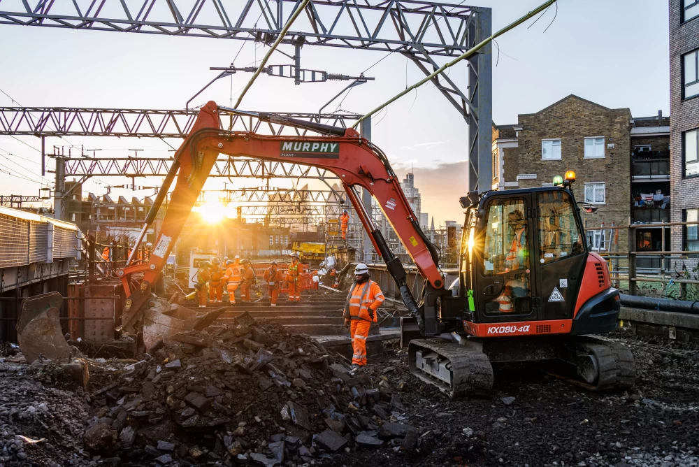 People working and operating machinery on a railway construction site with a cityscape in the background.