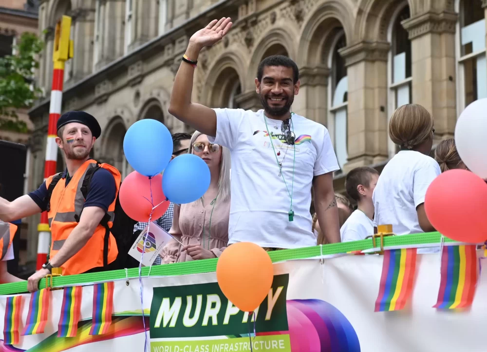 A man wearing a white Murphy t-shirt at a Pride event waving at the camera.