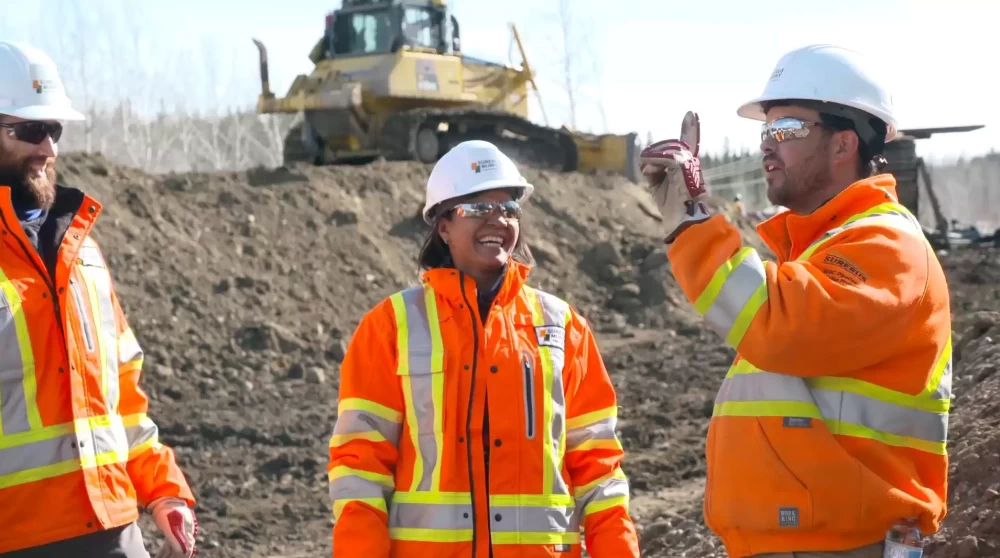 Three people, wearing Murphy high-visibility jackets and helmets, talking on a construction site with a construction vehicle in the background.