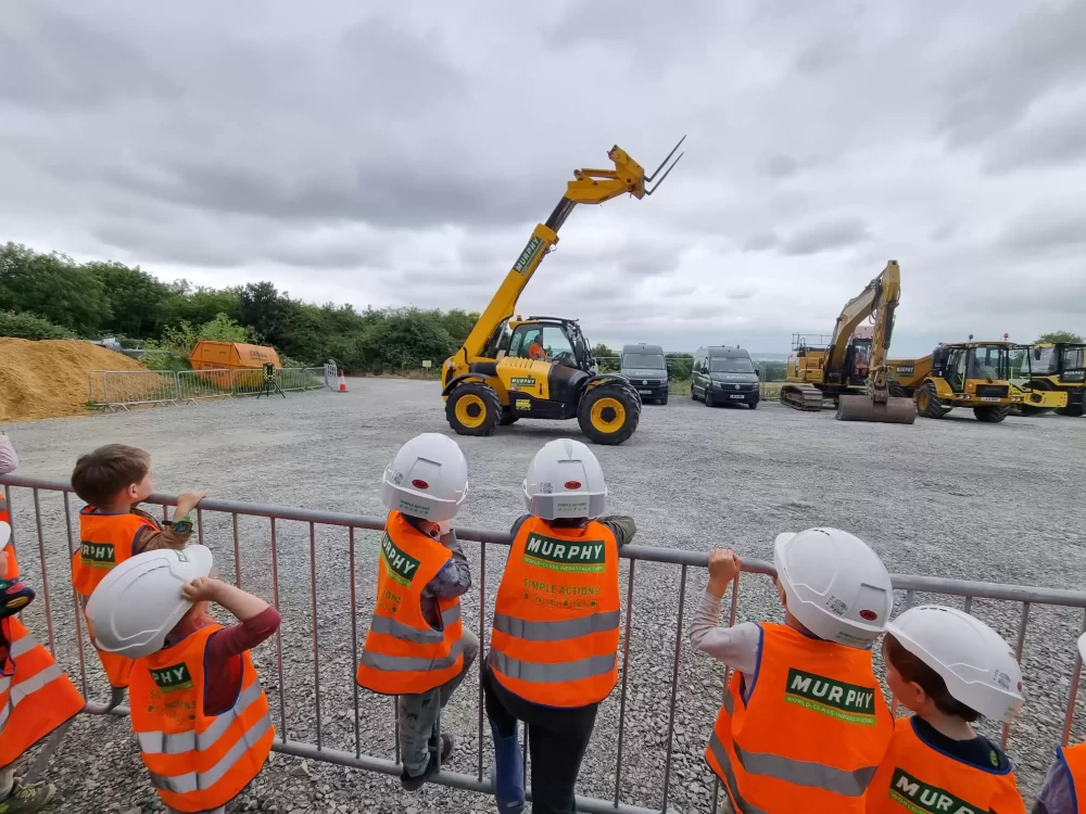 A group of children watching someone operate a construction vehicle from behind a metal barrier.