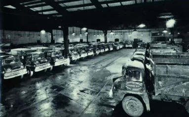 A black and white image of vehicles from the 1950s parked in a row in a warehouse.