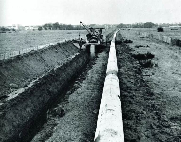 A black and white image of a construction vehicle creating a hole in the ground for a pipeline.