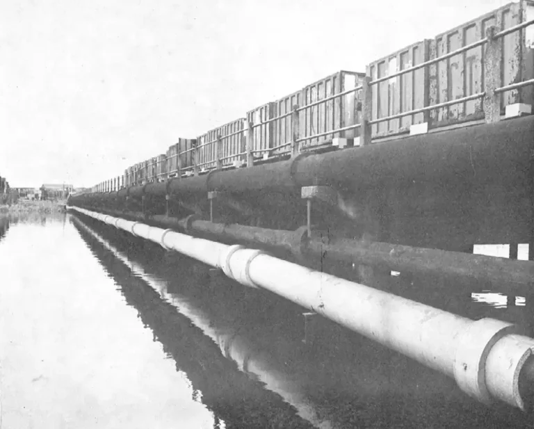 A black and white image of a river with a pipe and a metal containers in the background.