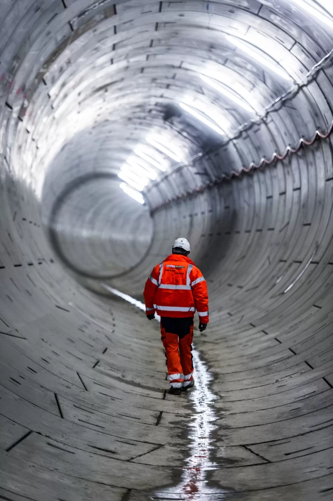 A person in orange safety uniform and a white helmet walking in a large concrete tunnel.