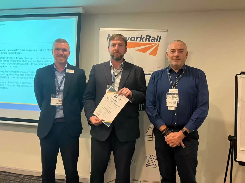 Three people, one holding a certificate, stood smiling in front of a projector screen and a Network Rail roller banner.