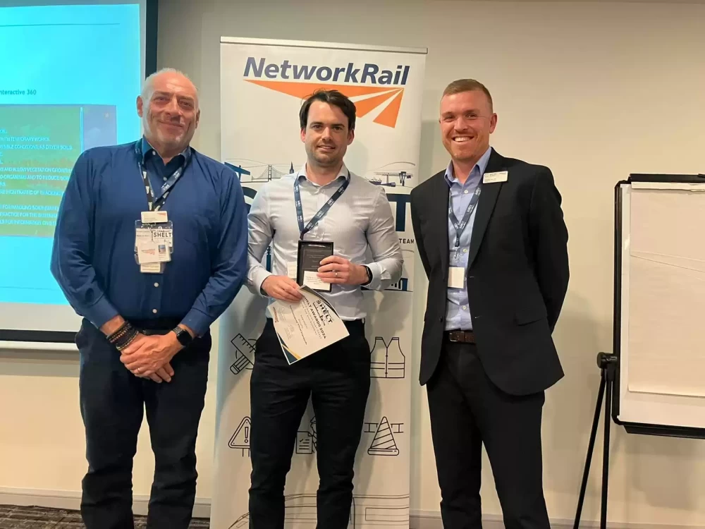 Three people in business dress stood smiling with a projector screen and a Network Rail roller banner in the background.