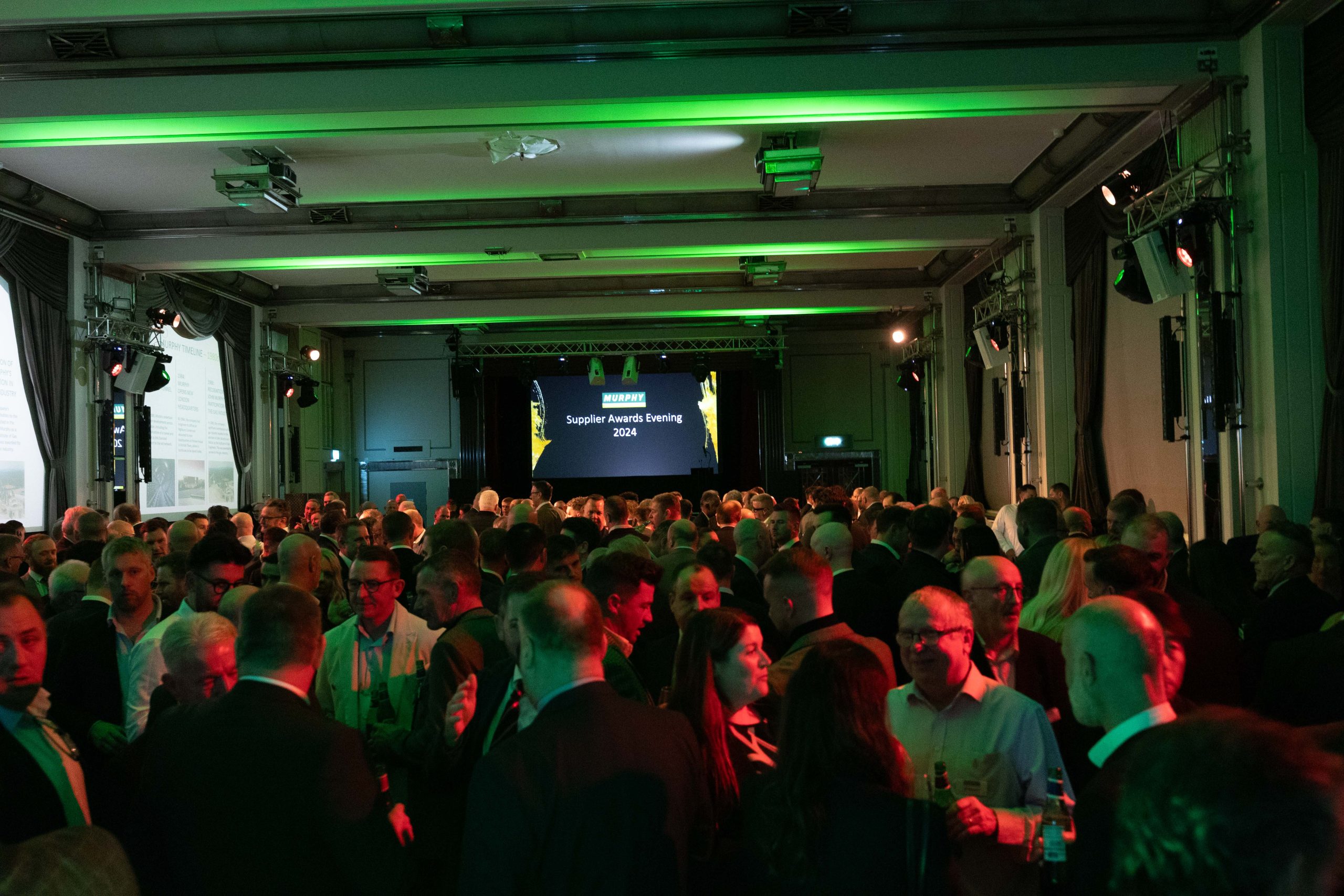 A dimly lit room full of people speaking to each other with a projector screen in the background.