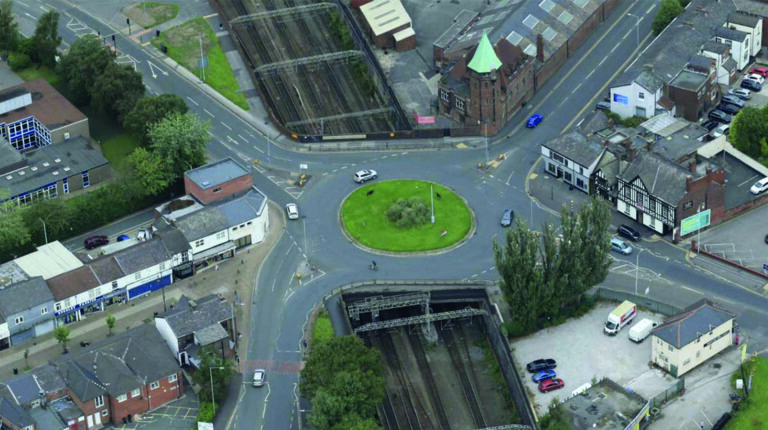 An aerial photo of a urban area in the daytime including a roads, a roundabout, cars, and train tracks.