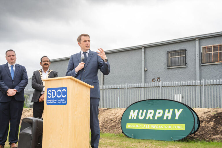 A person dressed in a suit giving a speech stood at a podium with the Murphy logo and a grey building in the background.