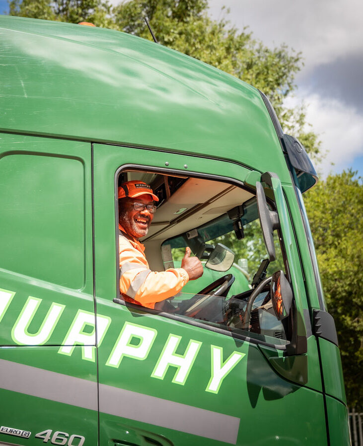 A person wearing a high-visibility uniform sat in the drivers seat of a murphy lorry smiling with their thumb up.