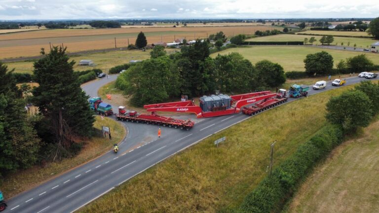 An aerial view of a construction vehicle moving heavy machinery on a road surrounded by fields.