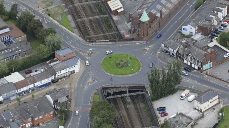 An aerial photo of a urban area in the daytime including a roads, a roundabout, cars, and train tracks.