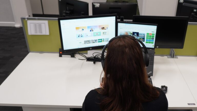 A person with headphones on sat at a desk looking at a laptop.