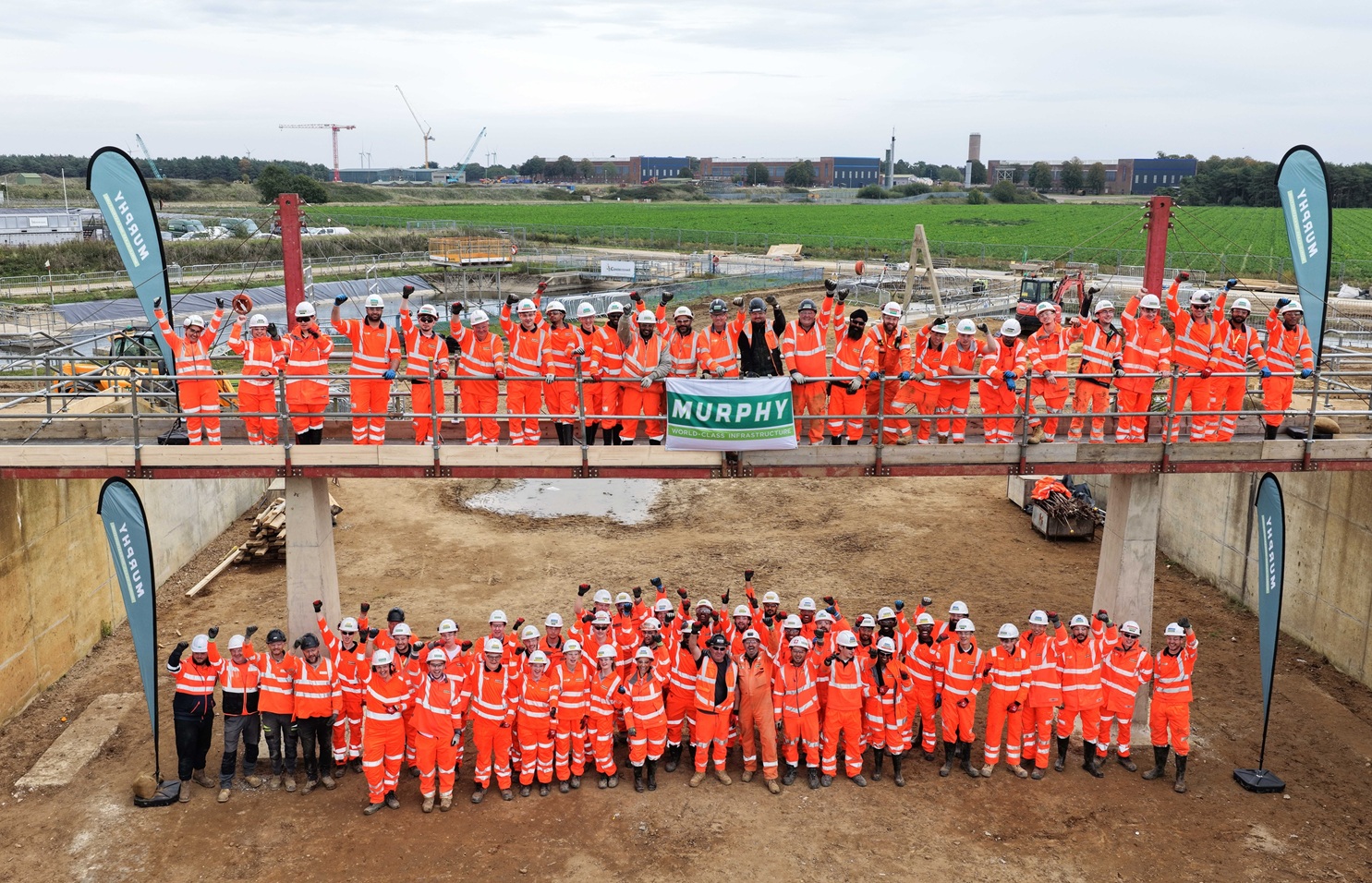 A large group of Murphy employees dressed in safety uniform with their arms raised at a construction site.