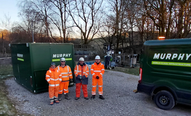 Four Murphy employees in orange high-visibility uniforms and white helmets stood on a construction site in front of a Murphy branded container and next to a Murphy van.