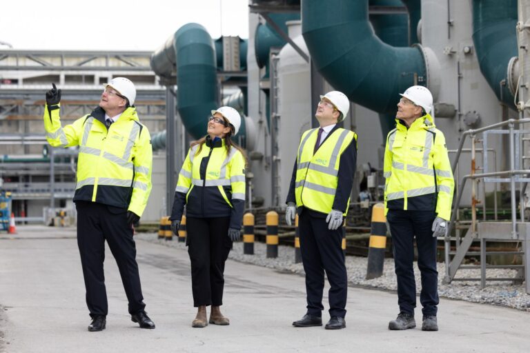 Four people stood in row in yellow high-visibility jackets, white helmets and safety glasses looking up.