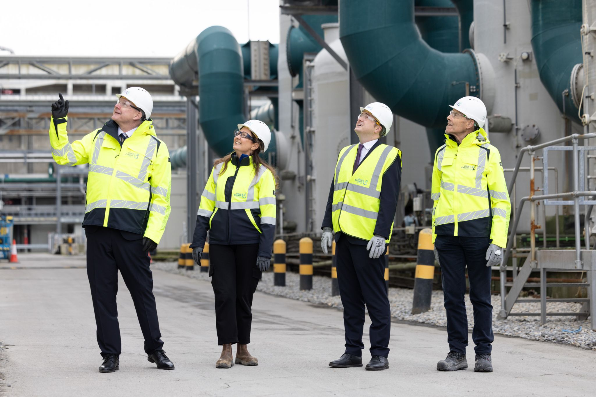 Four people stood in row in yellow high-visibility jackets, white helmets and safety glasses looking up.