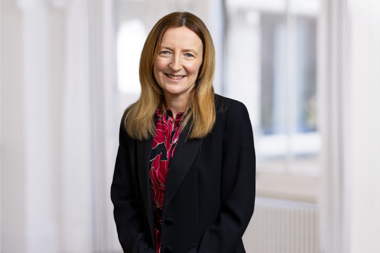 A woman wearing formal business wear smiling in a bright room with a window in the background.