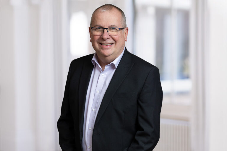 A man in business dress stood smiling stood in a white room with a window in the background