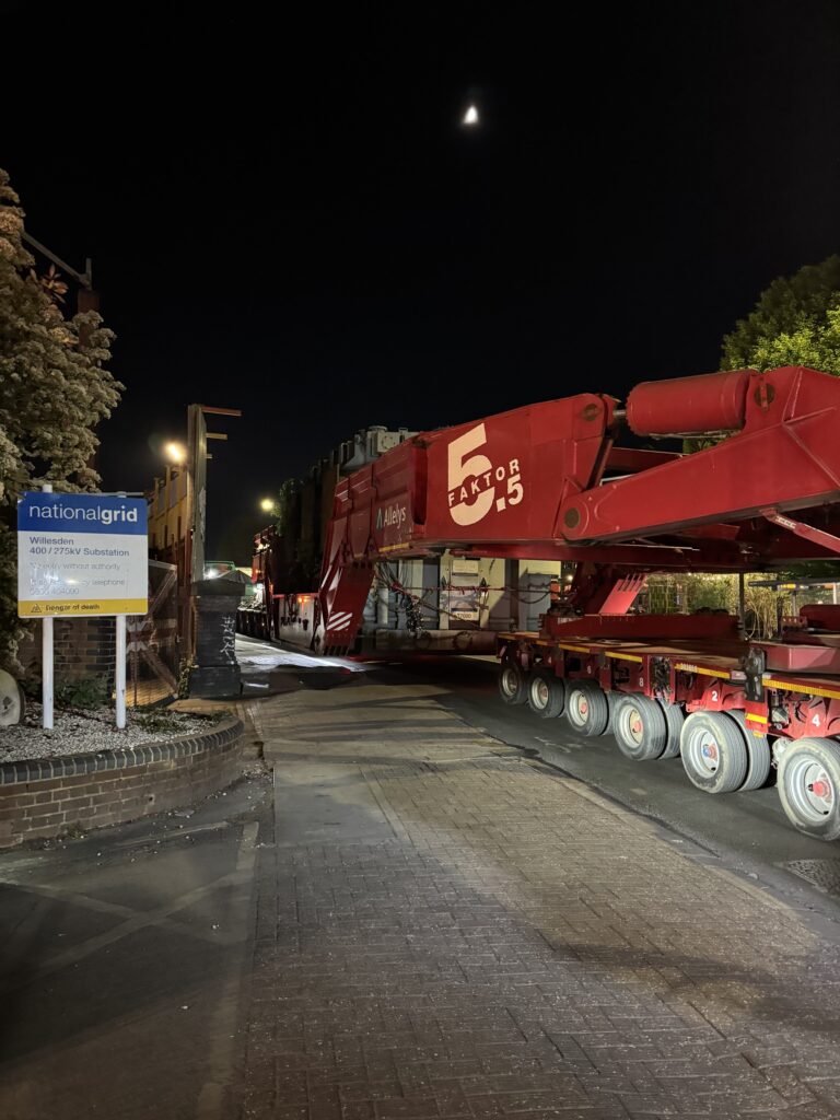 A large red transporter carrying a grey electrical transformer on a road at night time